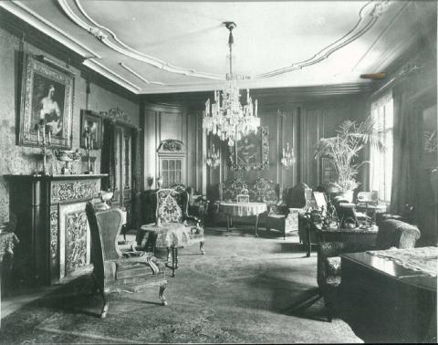 Blick in ein großbürgerliches Wohnzimmer mit Kamin, Luster, Gemälden und Klavier, Schwarz-Weiß-Foto