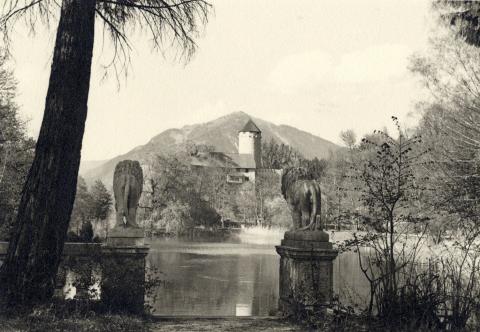 Blick über einen Teich auf das Schloss, im Vordergrund zwei steinerne Löwen auf Sockeln, Schwarz-Weiß-Foto